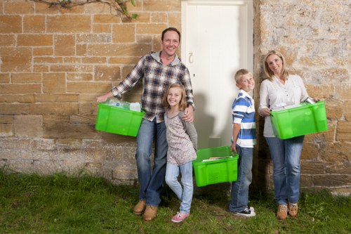 A family of four standing outdoors against an aged stone and brick wall, each person holding a bright green plastic rubbish bin. The father, dressed in a checkered shirt and jeans, stands on the left with a smile, holding a bin in his right hand. Next to him, a young girl with long blond hair, wearing a grey top and light jeans, also holds a bin in her right hand, smiling towards the camera. To her right, a boy in a striped blue and white shirt and dark jeans stands slightly behind the woman, who has shoulder-length blond hair, a white blouse, and jeans, and is holding a green bin in her left hand. The scene is set on a grassy patch with natural daylight, highlighting the textured wall behind them. This image illustrates a rubbish collection or waste disposal activity typical of residential waste management services, such as those offered by Commercial Waste Anerley in the Anerley area near London, with the family preparing for rubbish removal or disposal in a domestic setting.