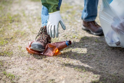 A person wearing a white work glove is seen picking up a discarded brown glass bottle from a patch of bare soil or gravel ground. The bottle, which appears to be an empty beer or soft drink container, has a blue cap and is lying on its side. The individual is wearing blue denim jeans and brown work boots, with part of a green jacket visible. Nearby, a large transparent plastic rubbish bag is partially visible on the right side of the image. The setting appears to be an outdoor area, possibly a driveway or a garden space, where rubbish such as bottles has been discarded. The background shows a blurred outdoor environment with natural light, indicating daytime, and the scene reflects an initiative of rubbish collection or litter picking, potentially by companies like Commercial Waste Anerley servicing the Anerley area with waste removal services.