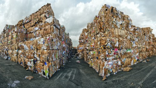 A large outdoor rubbish dump consisting of two massive stacks of compacted cardboard and paper waste piled high on either side of a narrow pathway. The stacks are composed of flattened cardboard boxes, some with visible printed logos and colourful graphics, while others are plain brown. The surfaces of the stacks appear rough and uneven, with layers of crumpled and folded cardboard creating a textured appearance. The ground beneath the piles is a dark asphalt or tarmac surface, slightly weathered with small patches of loose debris. Overhead, the sky is cloudy with a heavy overcast, casting diffuse light on the scene. This image illustrating waste management highlights the scope of commercial rubbish disposal, such as that managed by Commercial Waste Anerley, which services local businesses in Anerley, London, ensuring proper handling and removal of cardboard and paper waste as part of their rubbish collection practices.